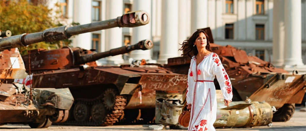 Woman in red and white dress walking by burned-out tanks in front of a large building with decorated columns