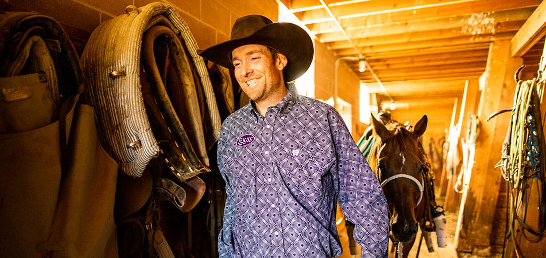 Smiling cowboy walking out of stable with horse following