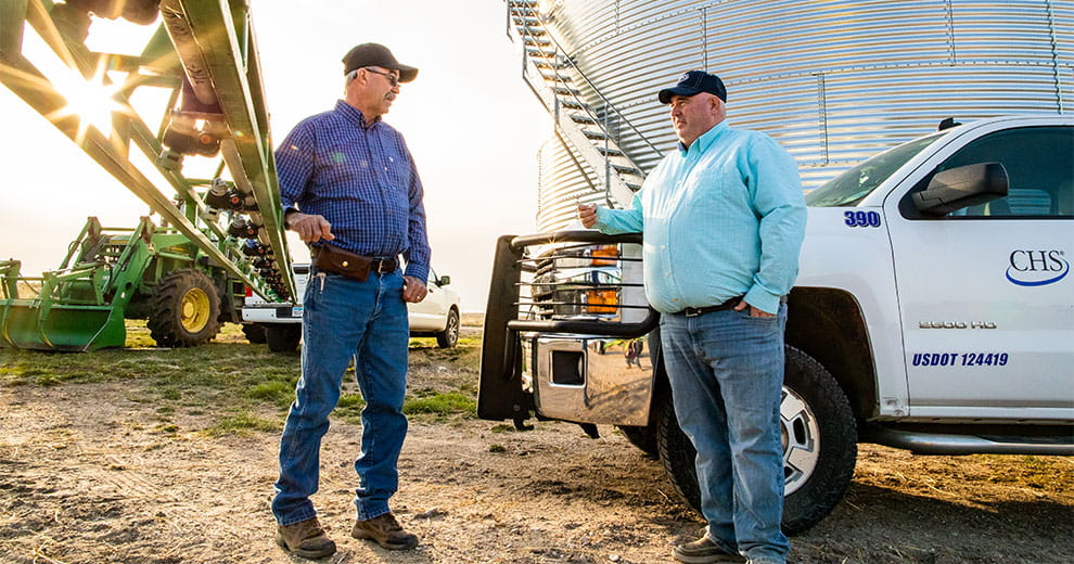 Two men standing beside a truck with a tractor and silo in the background