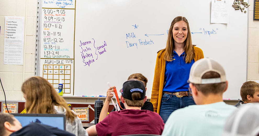 A teacher standing at the front of a classroom with a whiteboard behind her