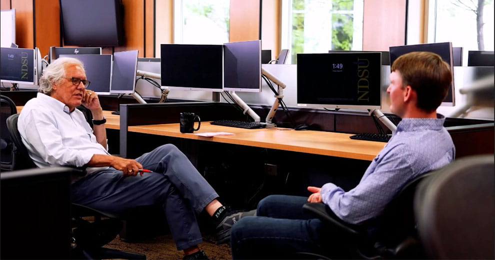 Professor and student sitting on chairs in front of computer monitors, facing each other and talking