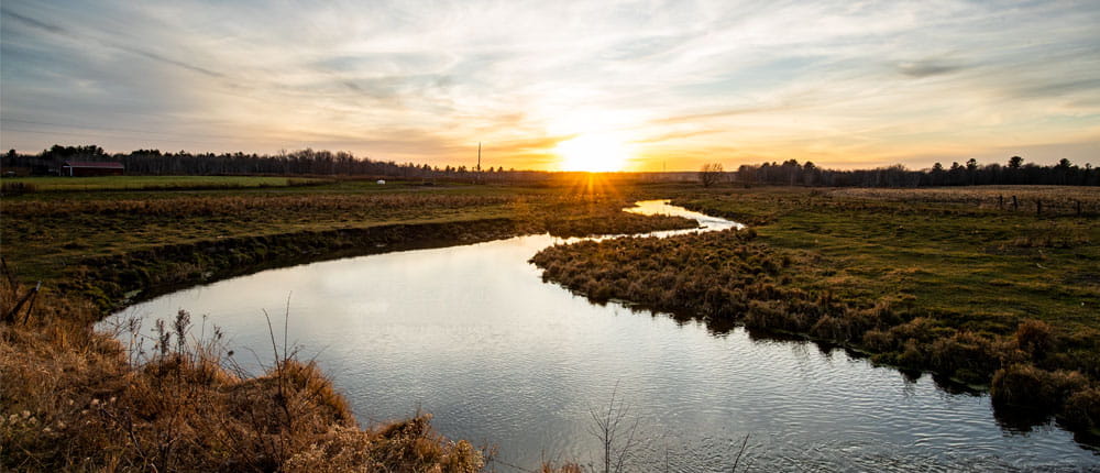 A river flows through a rural field with the sun setting in the distance.