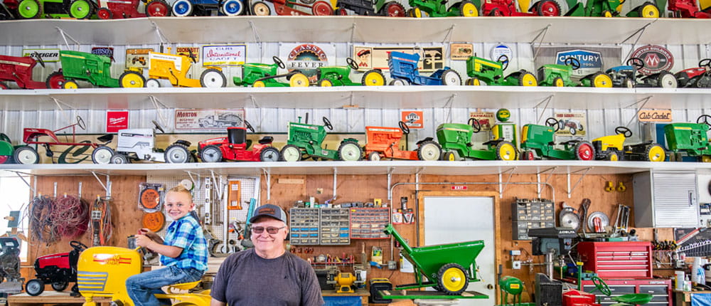 A man and his grandson pose in front of a wall of pedal tractors in a barn.