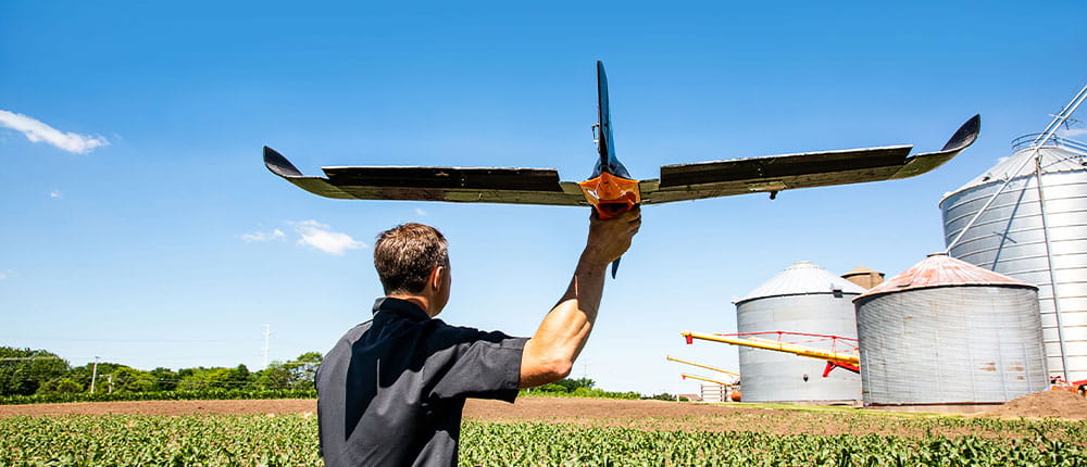 A man standing near a farm field prepares to launch a fixed-wing drone from his right hand