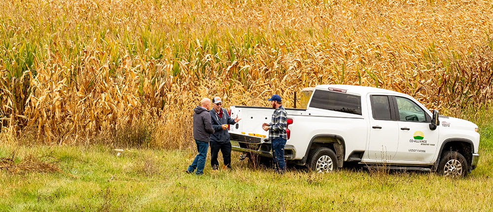 Three men talking by a truck with a Co-Alliance logo on the side, with a corn field in the background