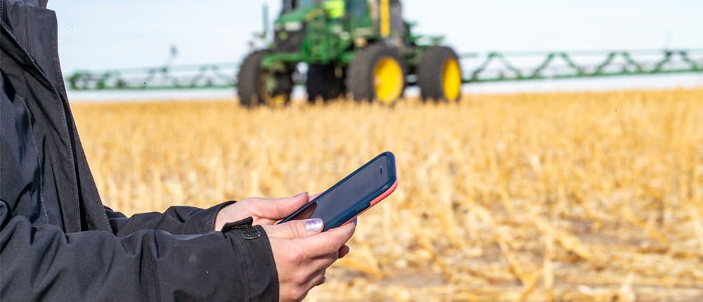 A farmer holds a phone in a farm field with a tractor in the background.