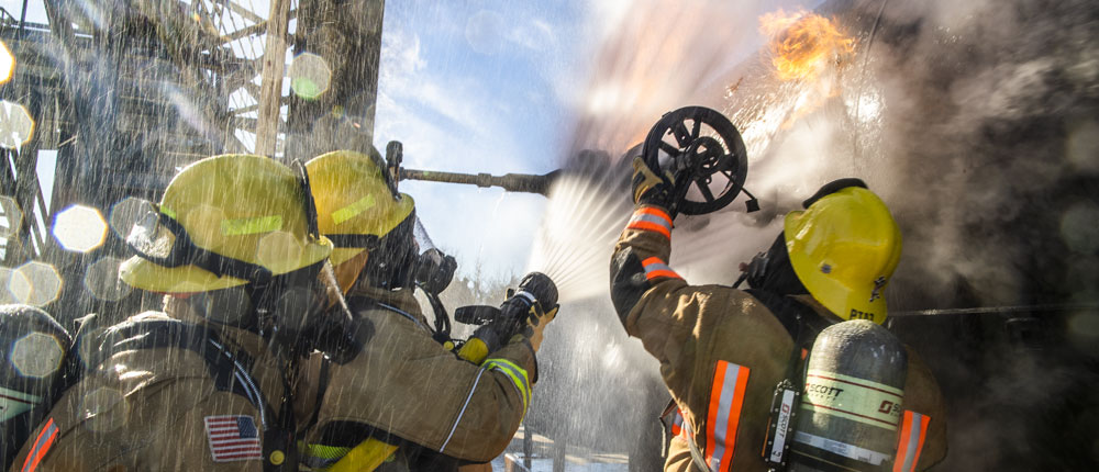 Three firefighters in safety gear use a water hose to put out a fire.