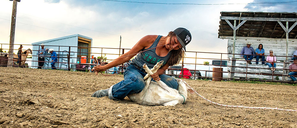 Teenager sits on top of goat that’s on the ground, typing its legs together with a rope