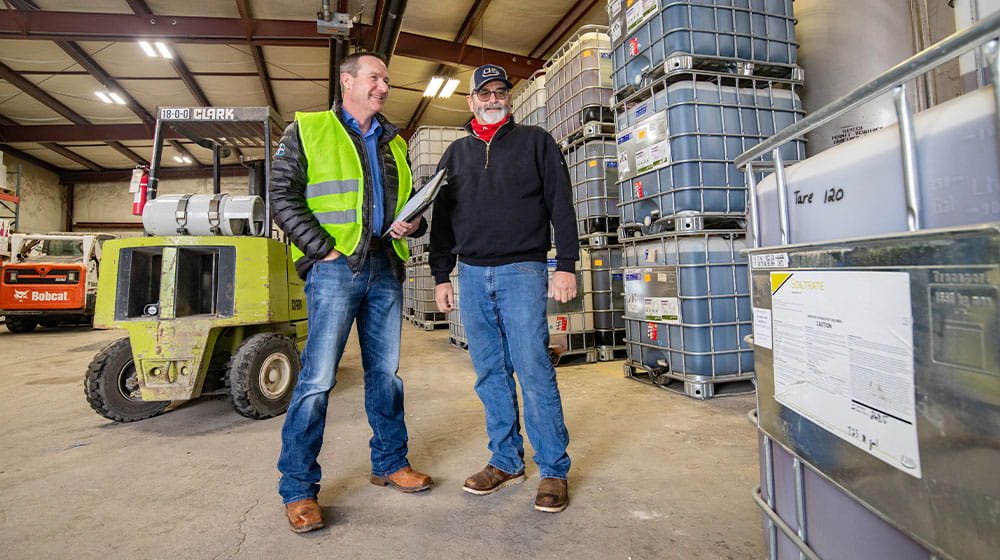 Two men standing in warehouse by herbicide totes